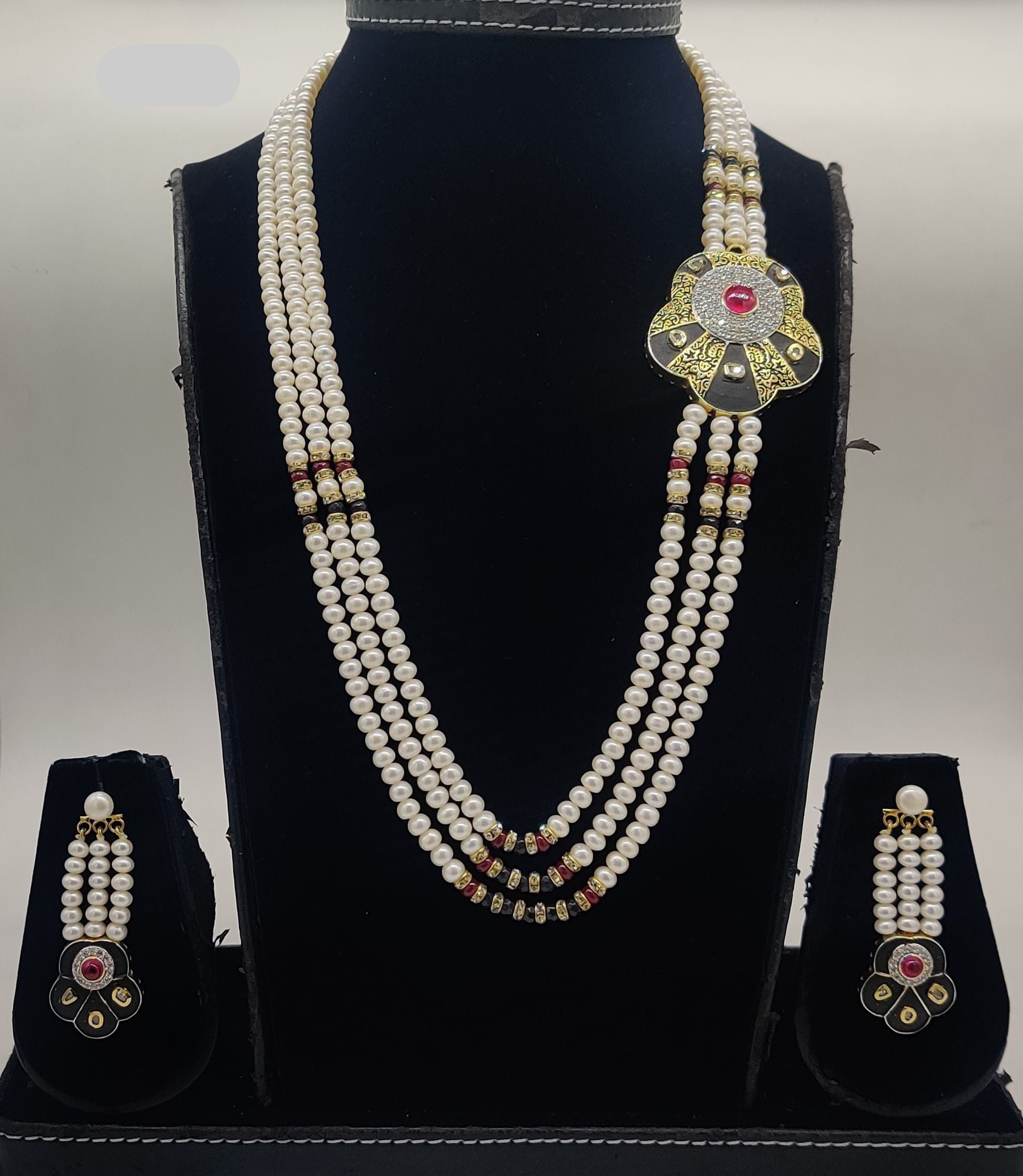 Three-line freshwater pearl broach set with black and gold Kundan centerpiece and red gemstone detailing with matching earrings.